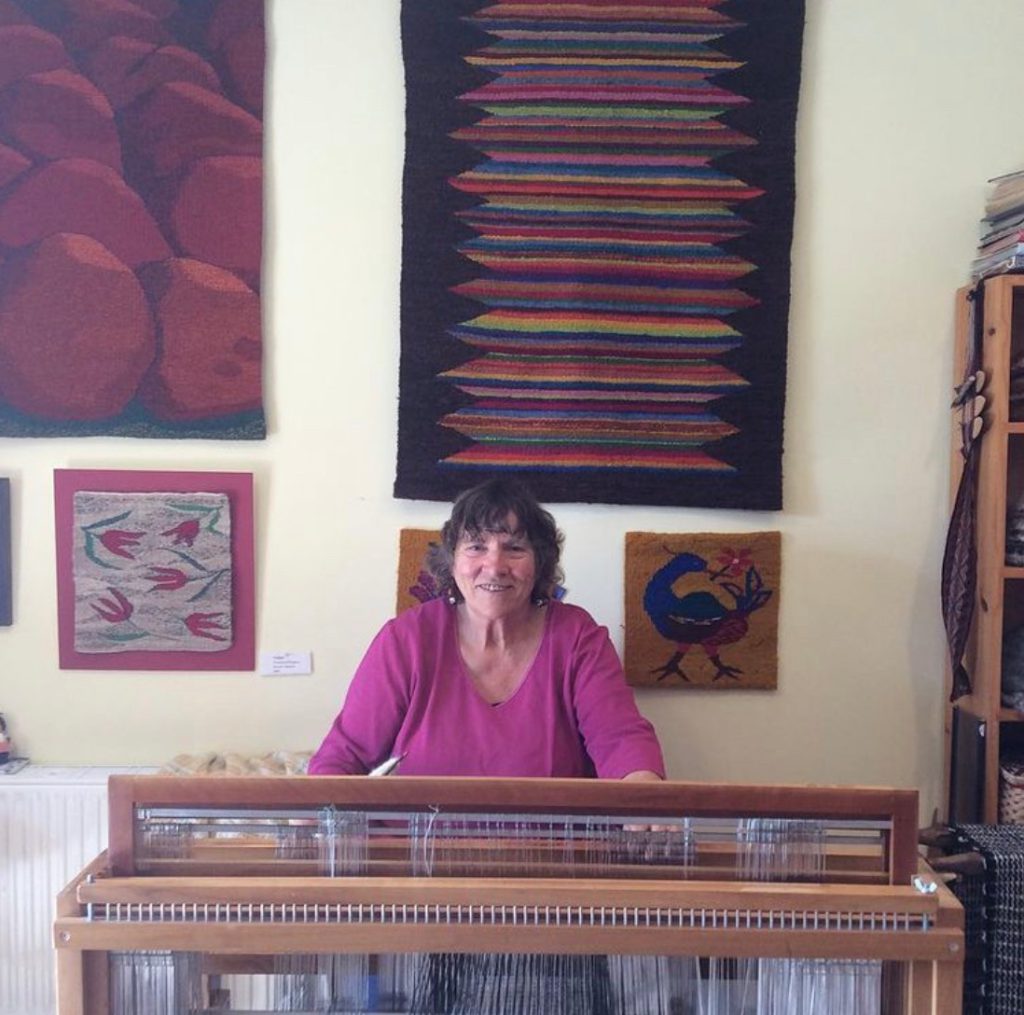 Annamaria at her loom in the studio, with her tapestries on the wall behind.