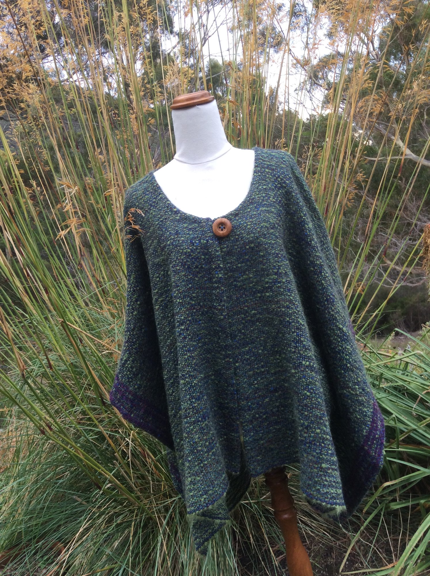 A handwoven cape in mossy greens and blues, with a purple border and a wooden button.
