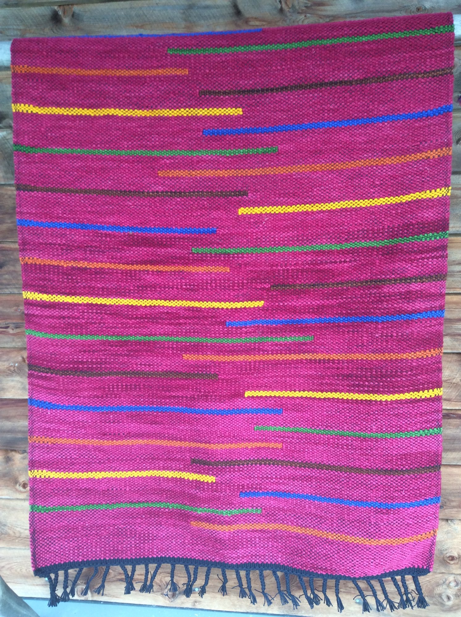 A floor rug in deep raspberry with stepped stripes of yellow, blue, green, orange and brown, finished with a black fringe.